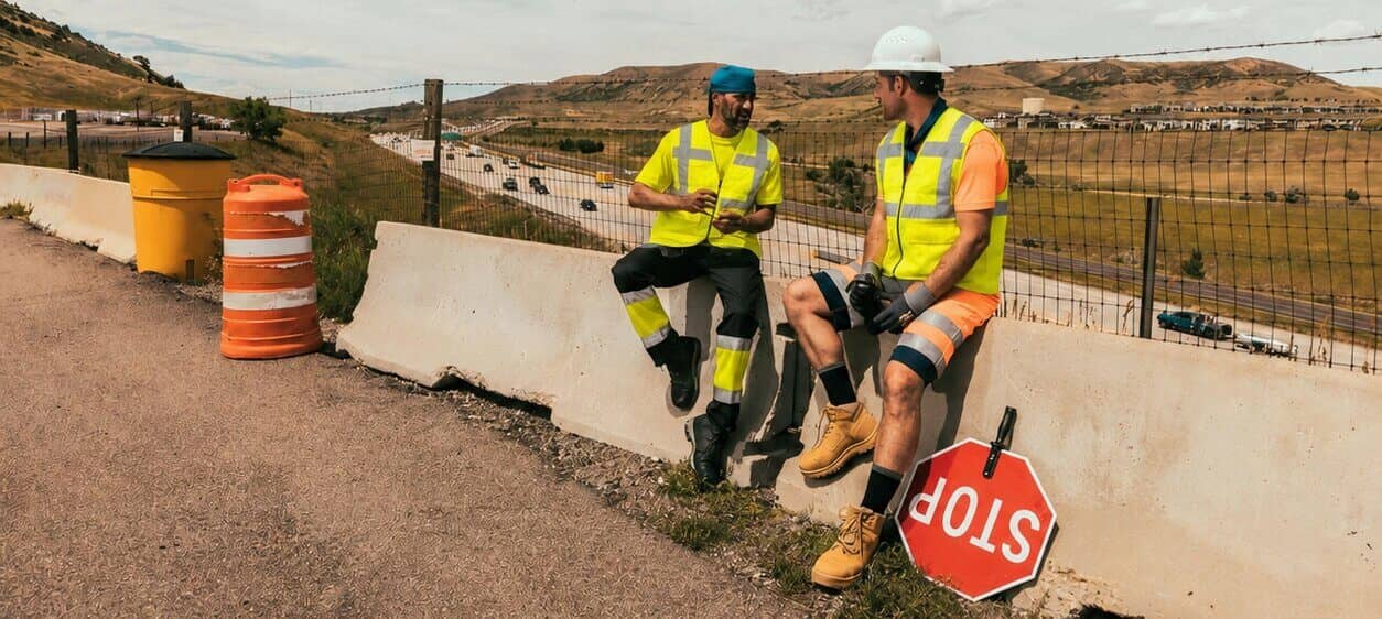 Dos trabajadores están sentados sobre una barrera de concreto al costado de la carretera. Llevan ropa de manga corta de alta visibilidad y chalecos refrigerantes. Al fondo, se aprecia un paisaje montañoso árido y un cielo azul nublado.