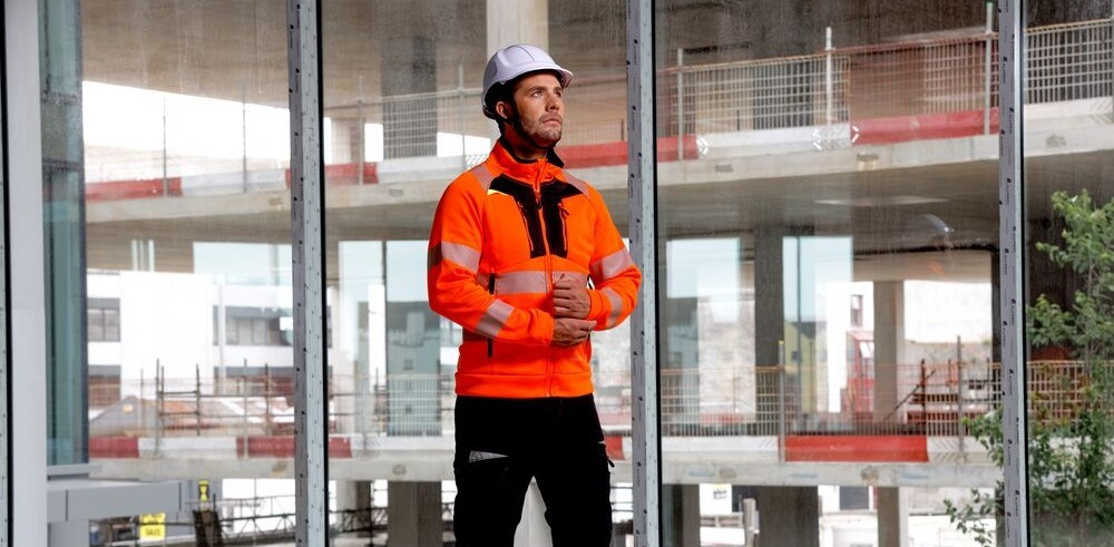 Un trabajador lleva la sudadera de alta visibilidad DX4 (modelo DX477). Viste un casco blanco y pantalones negros. Al fondo, se aprecian varios pisos de una casa en construcci&oacute;n a trav&eacute;s de una ventana de cuatro hojas.
