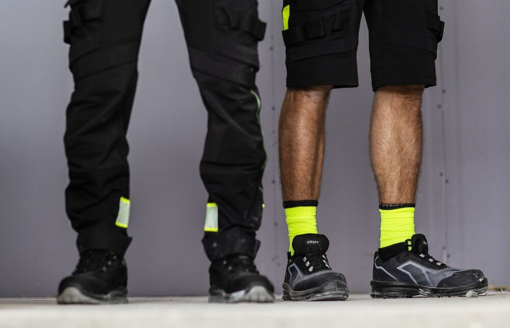 Se ven dos pares de piernas masculinas: un hombre lleva un pantal&oacute;n de trabajo largo negro y el otro un pantal&oacute;n corto de trabajo amarillo de alta visibilidad. Hay un enlace a nuestra categor&iacute;a de pantalones de trabajo.