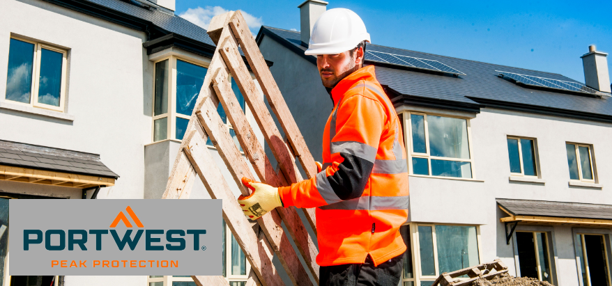 Trabajador con casco de seguridad blanco, guantes naranjas, pantalón negro y jersey naranja de alta visibilidad con franjas reflectantes. Al fondo se puede ver una hilera de casas casi completamente construidas con paredes pintadas de blanco y tejados oscuros. El cielo es azul, pasan algunas nubes blancas.