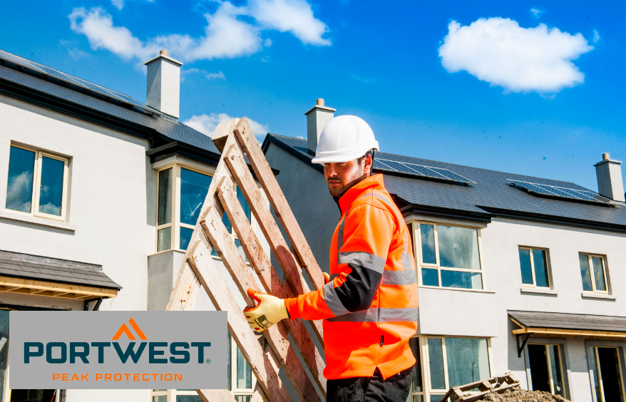 Trabajador con casco de seguridad blanco, guantes naranjas, pantalón negro y jersey naranja de alta visibilidad con franjas reflectantes. Al fondo se puede ver una hilera de casas casi completamente construidas con paredes pintadas de blanco y tejados oscuros. El cielo es azul, pasan algunas nubes blancas. Hay un enlace a nuestros suéteres de trabajo.