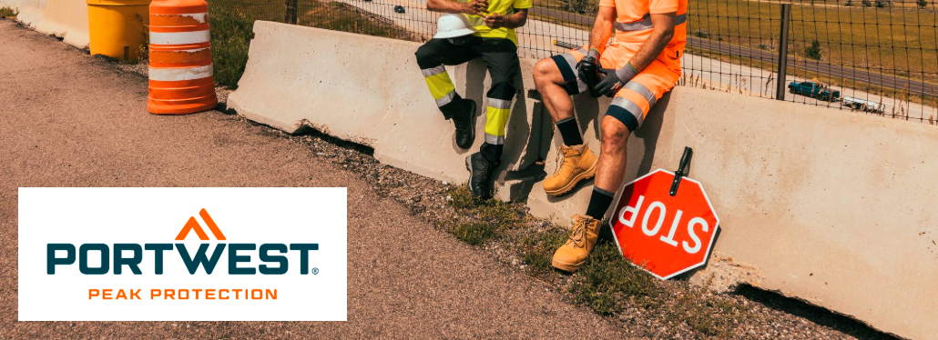 Se pueden ver las piernas de dos personas con ropa de trabajo. Estás sentado sobre una barrera de hormigón, a tu lado hay una señal de stop y al fondo hay una valla, una carretera y una cadena de colinas. El logotipo del fabricante Portwest en naranja y azul sobre fondo blanco se encuentra en la esquina inferior izquierda de la imagen.