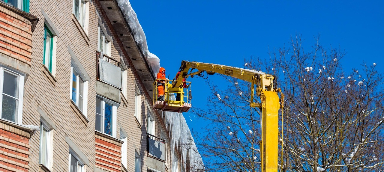 Una hilera de casas con car&aacute;mbanos en la cumbrera y nieve sobre el tejado. Un trabajador con ropa de alta visibilidad naranja est&aacute; de pie en una plataforma elevadora. A la derecha, se ve un &aacute;rbol desnudo con nieve en sus ramas.