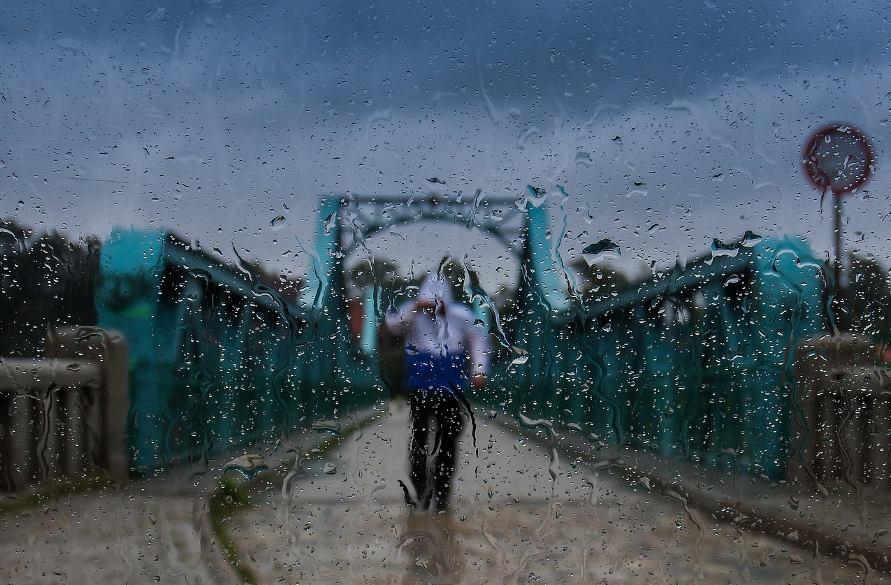 Puente azul contra un cielo gris con una persona con ropa de lluvia en medio de la carretera. La escena fue fotografiada a través de un panel de vidrio mojado. Se proporciona un enlace a toda nuestra selección de ropa impermeable práctica.