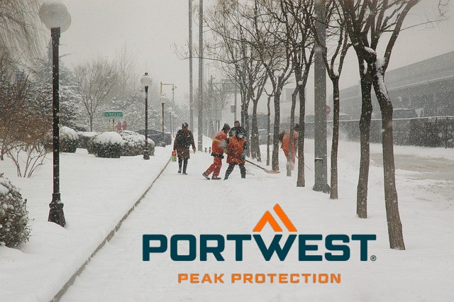 Acera cubierta de nieve con peatones muy abrigados, árboles y personas con ropa de alta visibilidad paleando nieve. Enlace a nuestra colección de invierno.
