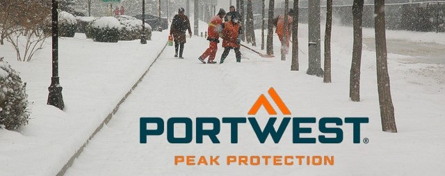 Acera cubierta de nieve con peatones muy abrigados, árboles y personas con ropa de alta visibilidad paleando nieve.