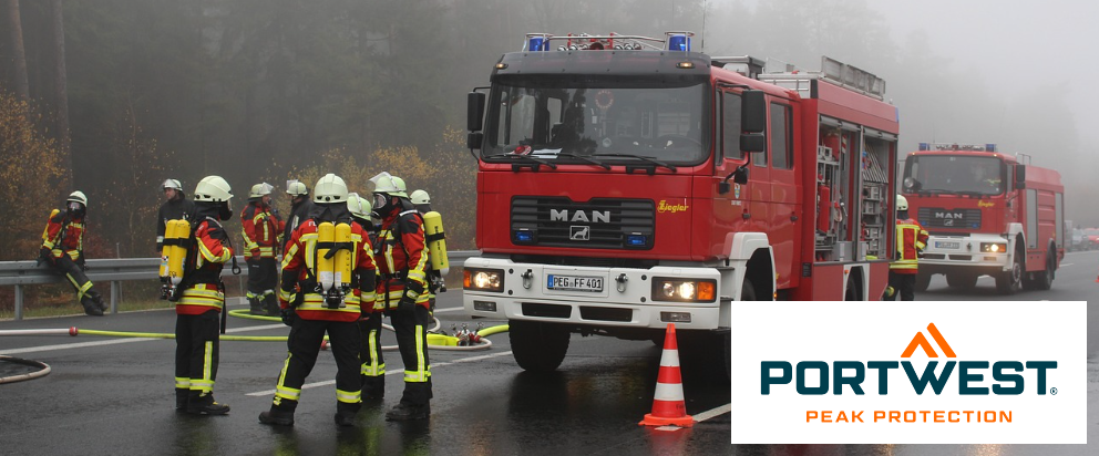 Escena callejera con dos camiones de bomberos rojos y varios bomberos. Al fondo se pueden ver árboles cubiertos de niebla. En la esquina inferior derecha está el logotipo de Portwest en color azul anaranjado delante de un rectángulo blanco.