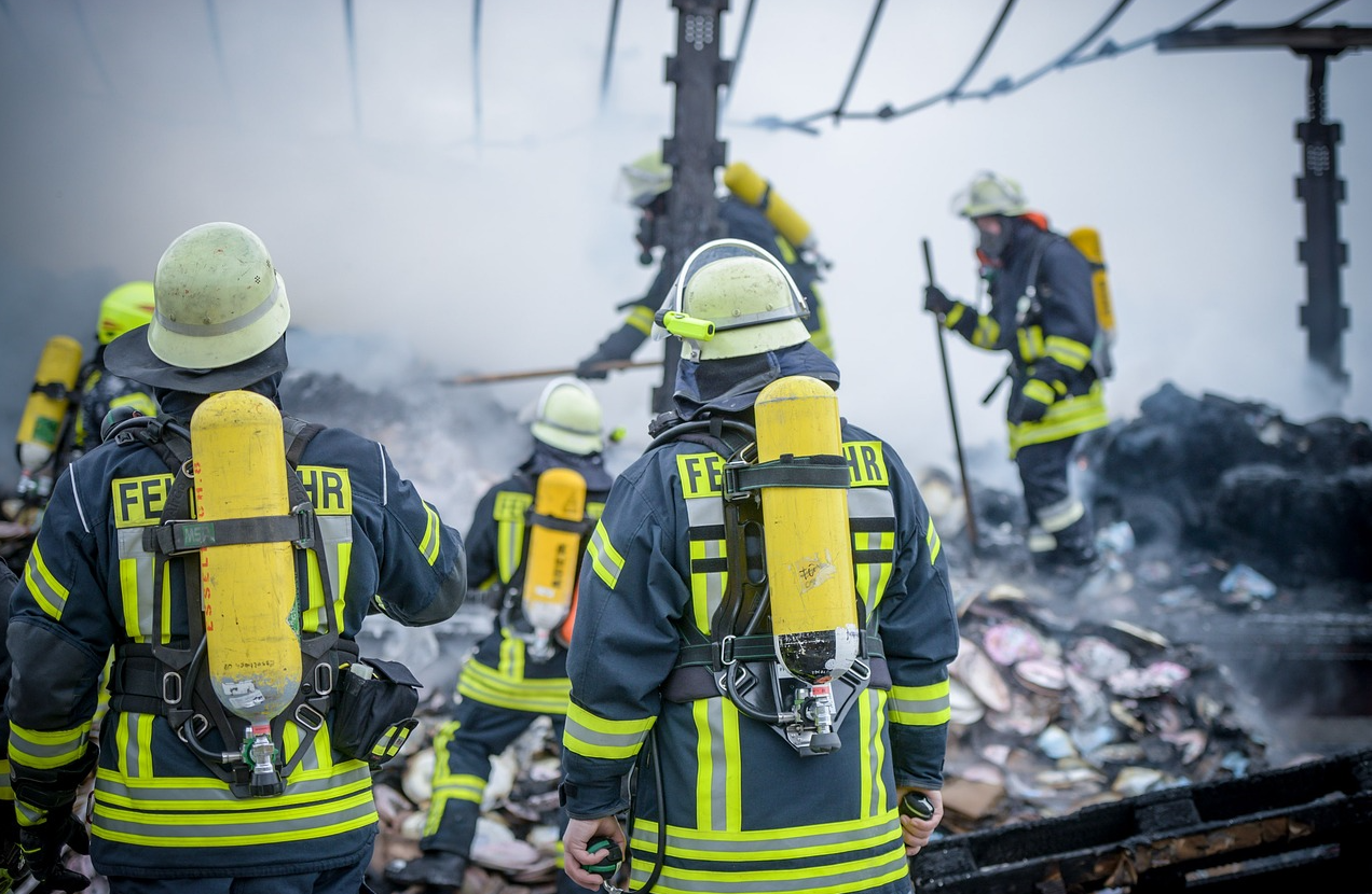 Bomberos vestidos de azul con franjas amarillas de advertencia y cascos en un depósito de chatarra. El enlace proporcionado conduce a nuestra selección de ropa de extinción de incendios ignífuga.