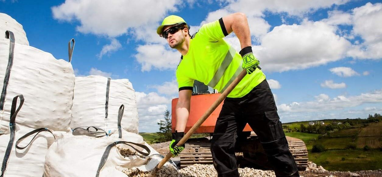 Un hombre con camisa amarilla y pantalón negro. Lleva una gorra amarilla y gafas de sol, y está paleando grava de grandes sacos. Al fondo, se aprecia un cielo azul ligeramente nublado, una obra en construcción y colinas verdes.