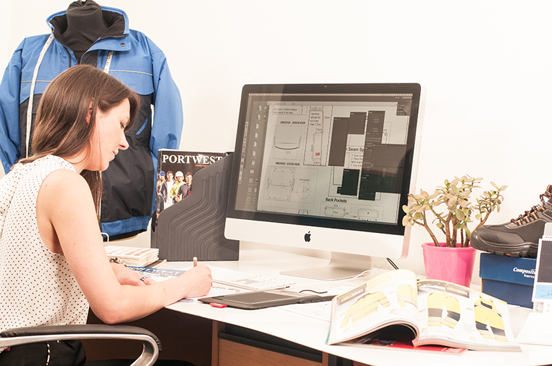 Mujer morena trabajando en el escritorio. Los dibujos técnicos de la ropa de trabajo se pueden encontrar en la pantalla de su computadora.