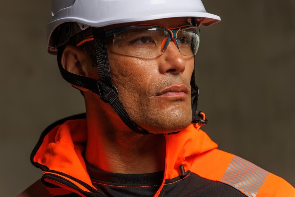Primer plano de un trabajador con casco de seguridad blanco y gafas de protección transparentes, mirando concentrado a lo lejos; lleva ropa de alta visibilidad naranja-negra. Enlace a nuestra colección de protección ocular incluido.