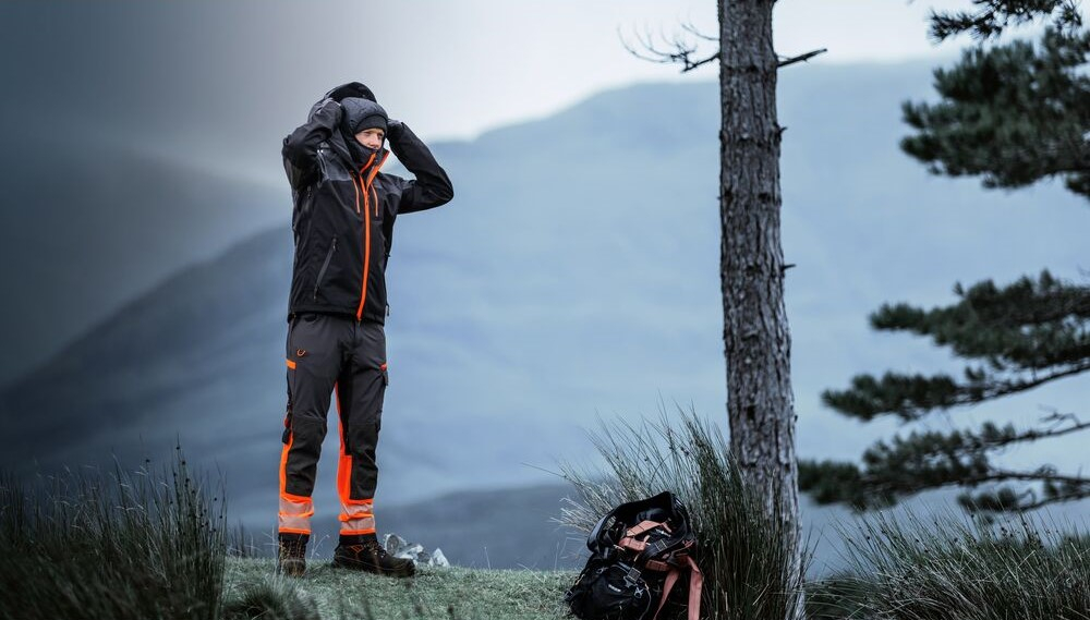 Trabajador con ropa de trabajo resistente al clima con elementos reflectantes está al aire libre en las montañas y se pone un gorro de invierno. Un enlace a nuestros gorros está incluido.