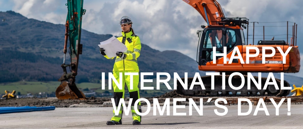 La imagen muestra a una mujer con equipo de seguridad de color amarillo brillante, que incluye casco y gafas protectoras, parada en un sitio de construcción sosteniendo planos. Detrás de ella hay una gran excavadora naranja con el brazo levantado, y al fondo se extienden colinas verdes bajo un cielo parcialmente nublado. Sobre la imagen está escrito en grandes letras blancas "HAPPY INTERNATIONAL WOMEN´S DAY!" en Inglés.