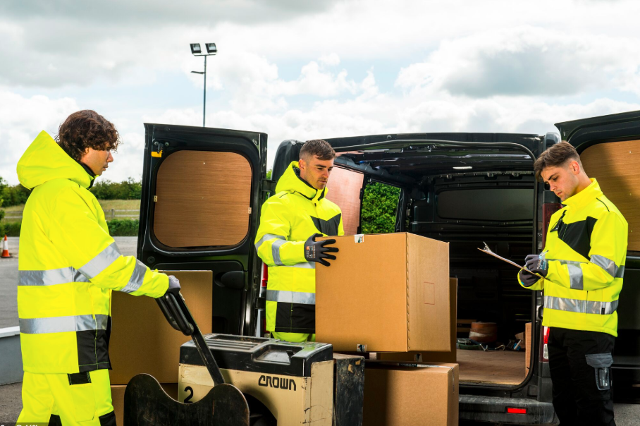 Tres hombres con chaquetas softshell amarillas de alta visibilidad con franjas reflectantes trabajan frente a una furgoneta abierta. Uno empuja un palé de cajas con una transpaleta, otro levanta una caja grande de la furgoneta y el tercero escribe algo en una carpeta. La escena es al aire libre bajo un cielo nublado. Hay un enlace a nuestras chaquetas softshell.