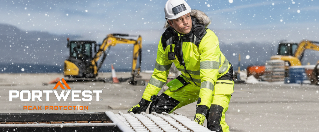 Un trabajador con ropa de invierno reflectante amarilla trabaja al aire libre bajo la nieve. Lleva casco y guantes. Al fondo se ven vehículos de construcción y un paisaje nevado. En la esquina inferior izquierda, el logotipo del fabricante Portwest aparece en letras blancas y naranjas.