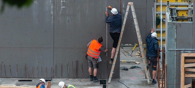 Se observa a dos trabajadores en una obra. Uno está de pie sobre una escalera y el otro la sujeta. Ambos llevan pantalones cortos de trabajo; uno lleva un casco blanco y el otro un chaleco reflectante naranja.