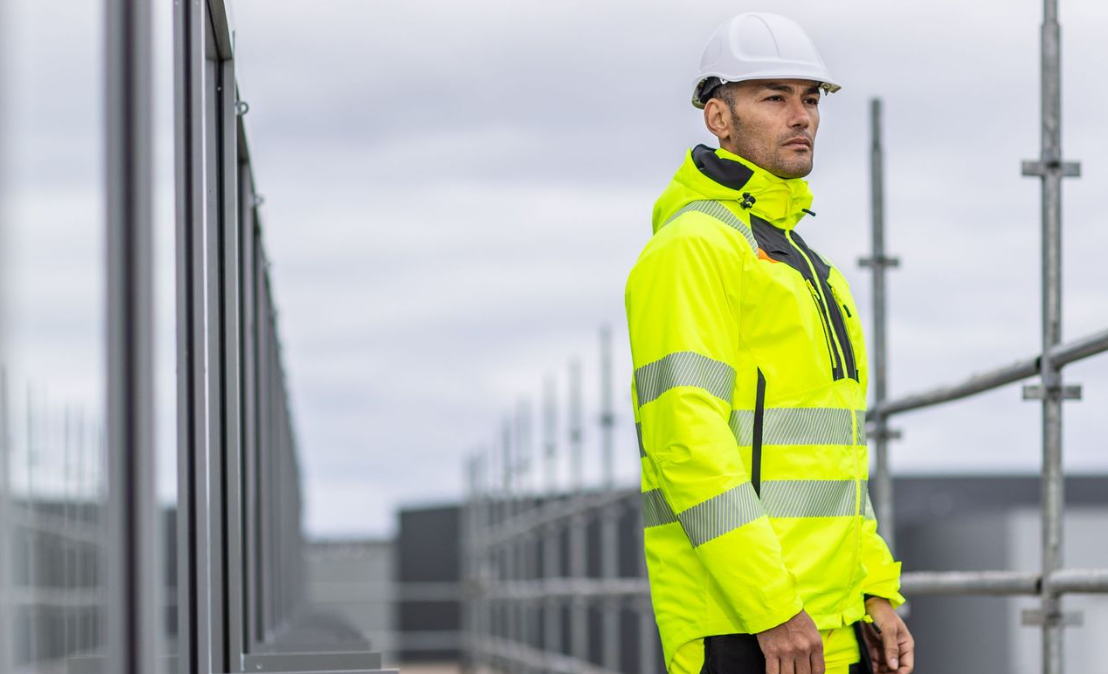 Un trabajador con ropa de trabajo amarilla de alta visibilidad y un casco blanco frente a un frente de ventana y la parte superior de un andamio vinculado a la colección DX4.