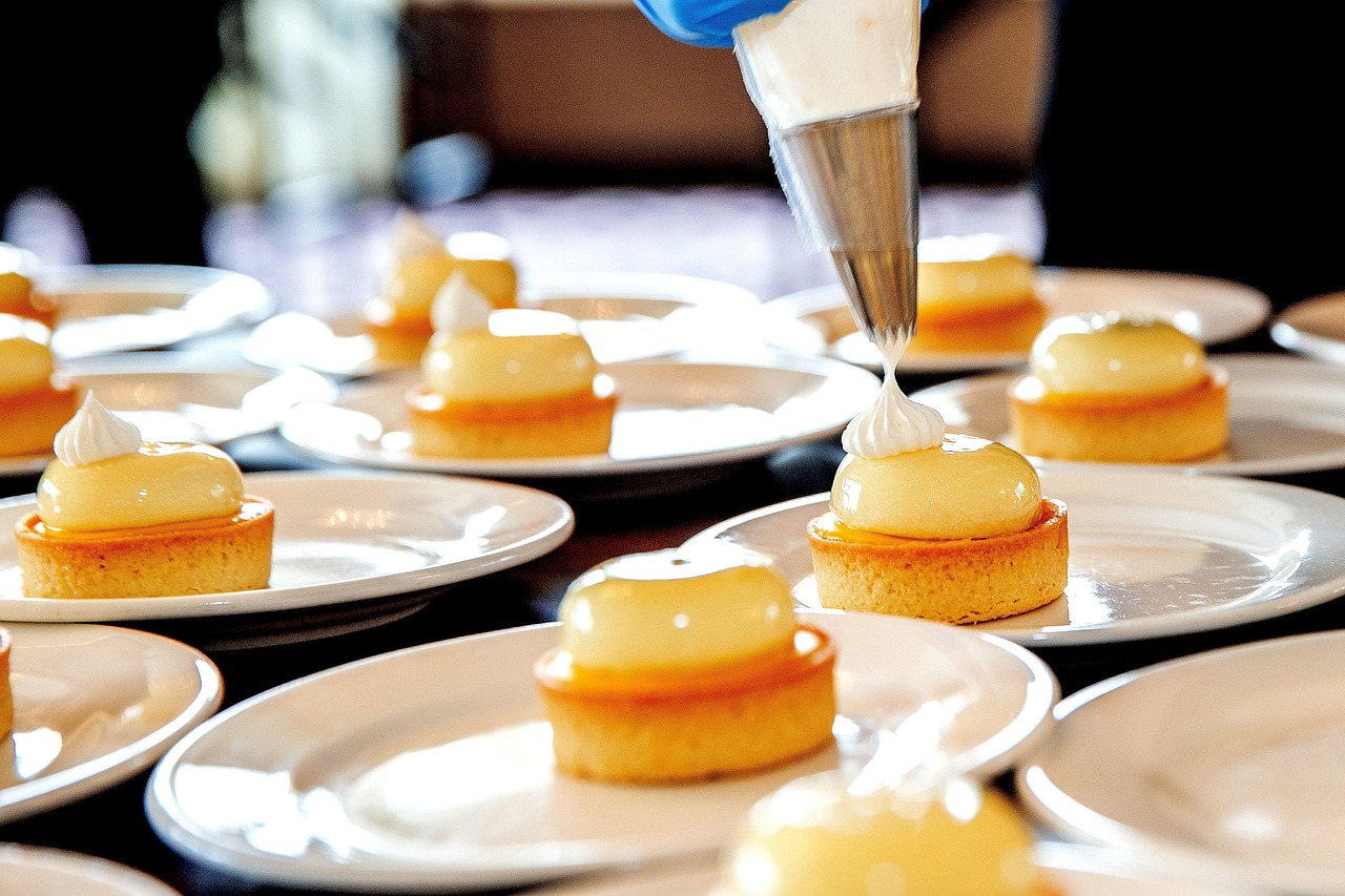Plato con reposter&iacute;a fina. Una mano con guante azul sostiene una manga pastelera y aplica una masa blanca sobre un peque&ntilde;o pastel.
