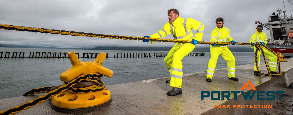 Tres trabajadores con ropa amarilla de alta visibilidad tirando de una cuerda. Al fondo se ve un barco, un puerto y una cadena montañosa nublada, en primer plano hay un bolardo amarillo. En la esquina inferior derecha de la imagen se encuentra el logotipo del fabricante Portwest en letras azules y naranjas.