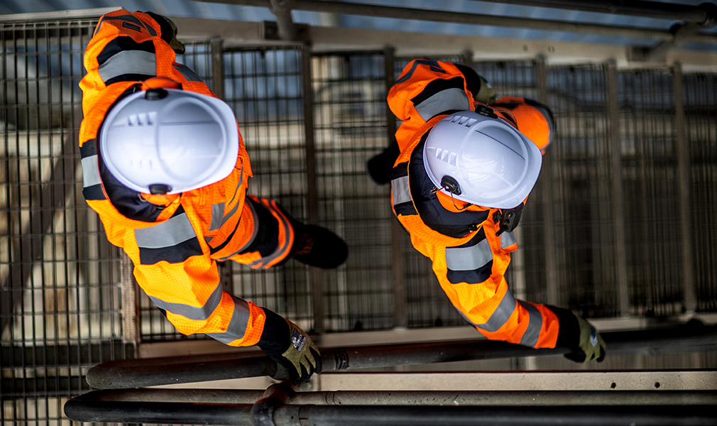 Vista aérea de dos trabajadores con cascos blancos, guantes negros y verdes, botas de seguridad y overoles naranjas de alta visibilidad con enlace a nuestra colección de ropa de protección contra arco eléctrico.