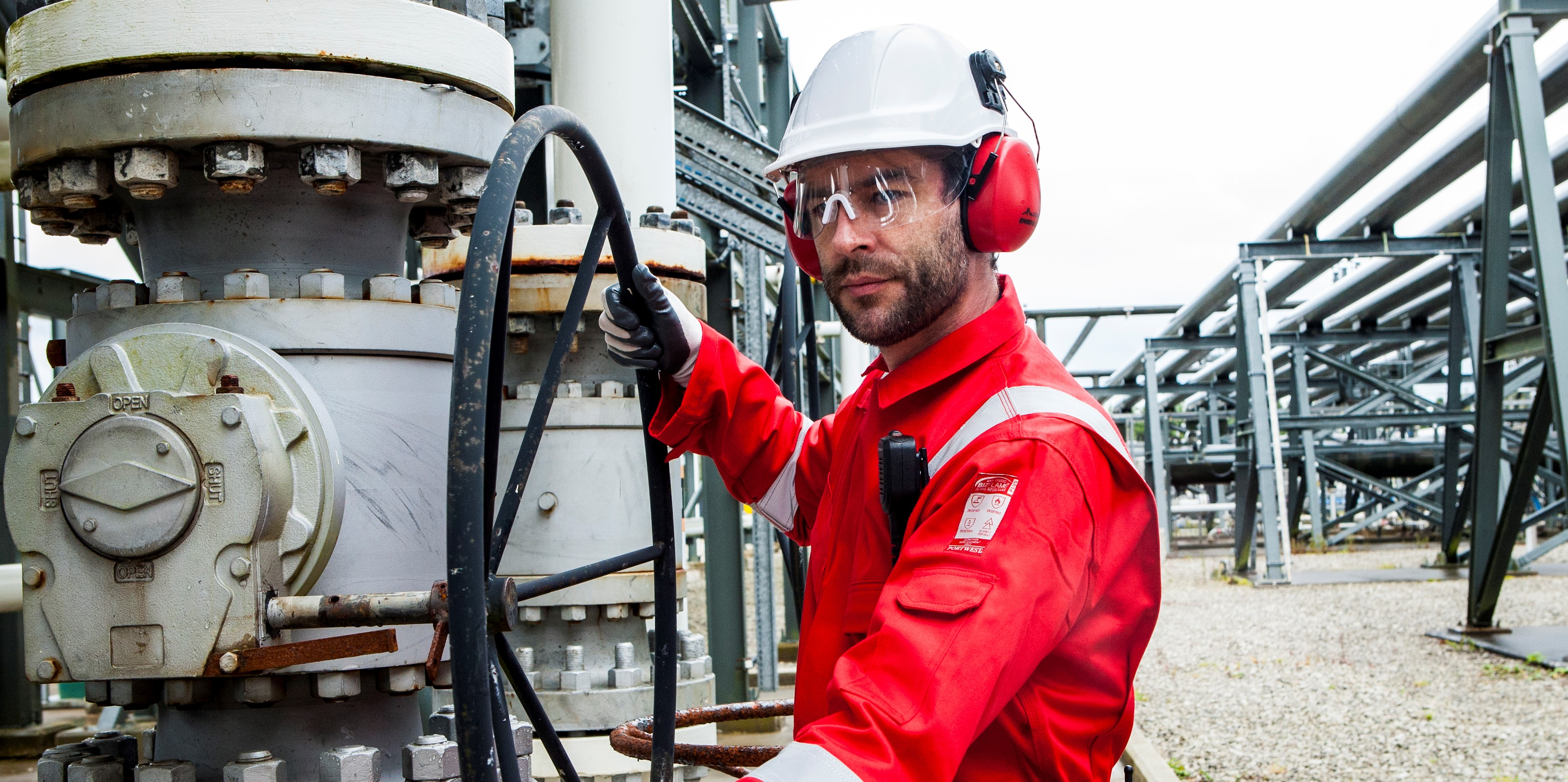 Trabajador en un contexto industrial con ropa de protección: casco blanco, gafas protectoras, orejeras rojas, guantes negros y mono rojo FR50 con tiras reflectantes.