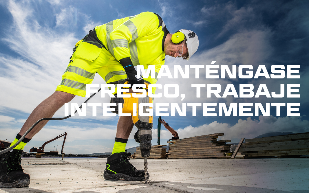 Trabajador con casco y protección auditiva en ropa de trabajo de verano amarilla de alta visibilidad trabajando con un martillo neumático, en el fondo azul, cielo ligeramente nublado, excavadora y pila de tablones de madera.