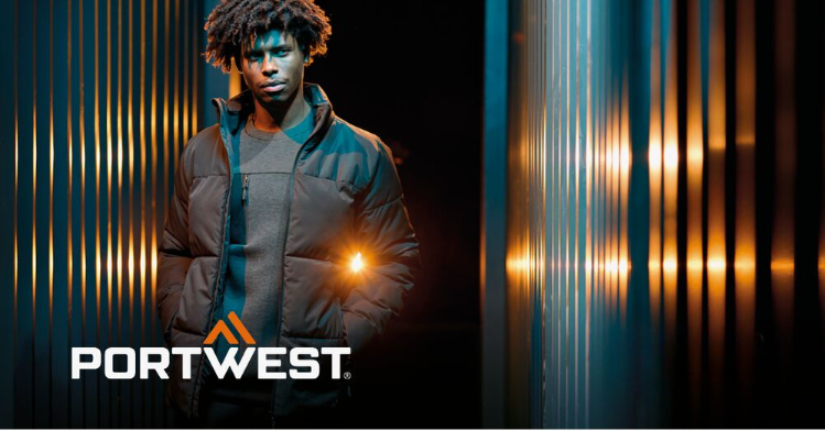 Un trabajador con ropa de trabajo negra sobre un fondo abstracto con una pared rayada y reflejos de luz. El logotipo del fabricante, Portwest, en blanco y naranja, se encuentra en la esquina inferior izquierda de la imagen.
