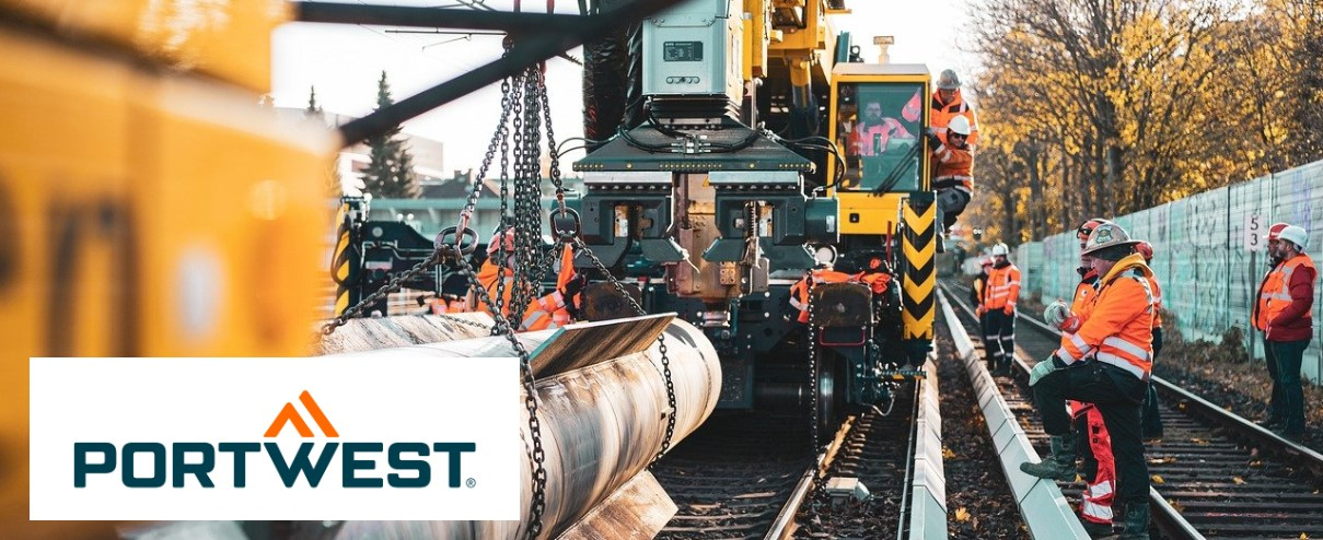 Varios trabajadores vestidos con ropa reflectante de color naranja y cascos de seguridad realizan trabajos de construcción en una línea ferroviaria. Al fondo se puede ver maquinaria pesada como una grúa y un vehículo ferroviario. Logotipo de Portwest visible en la esquina.