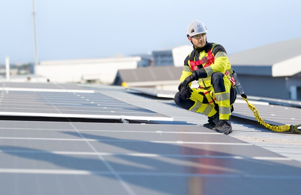 Trabajador en un tejado entre instalaciones fotovoltaicas. Lleva ropa de trabajo amarillo de advertencia, casco blanco, guantes y calzado negro. Está asegurado con el arnés Portwest 3-Punto Plus FP18. Enlace a nuestra protección contra caídas.