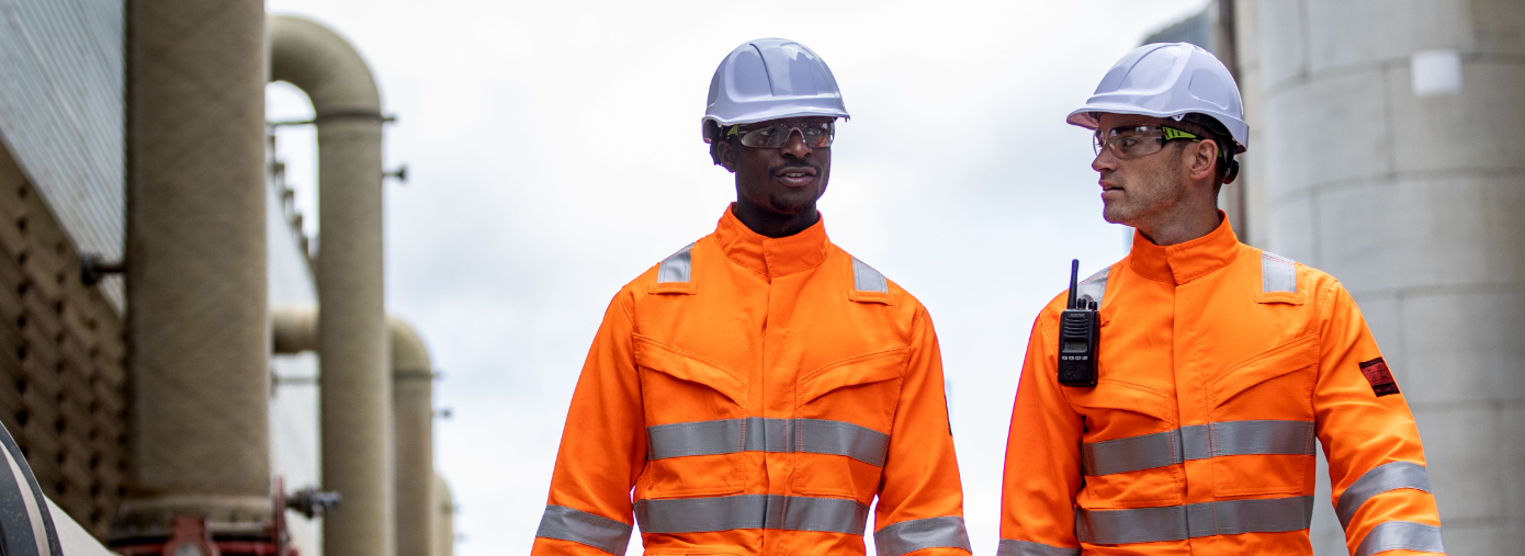 Dos trabajadores frente a un fondo industrial vistiendo ropa naranja de alta visibilidad y cascos blancos.