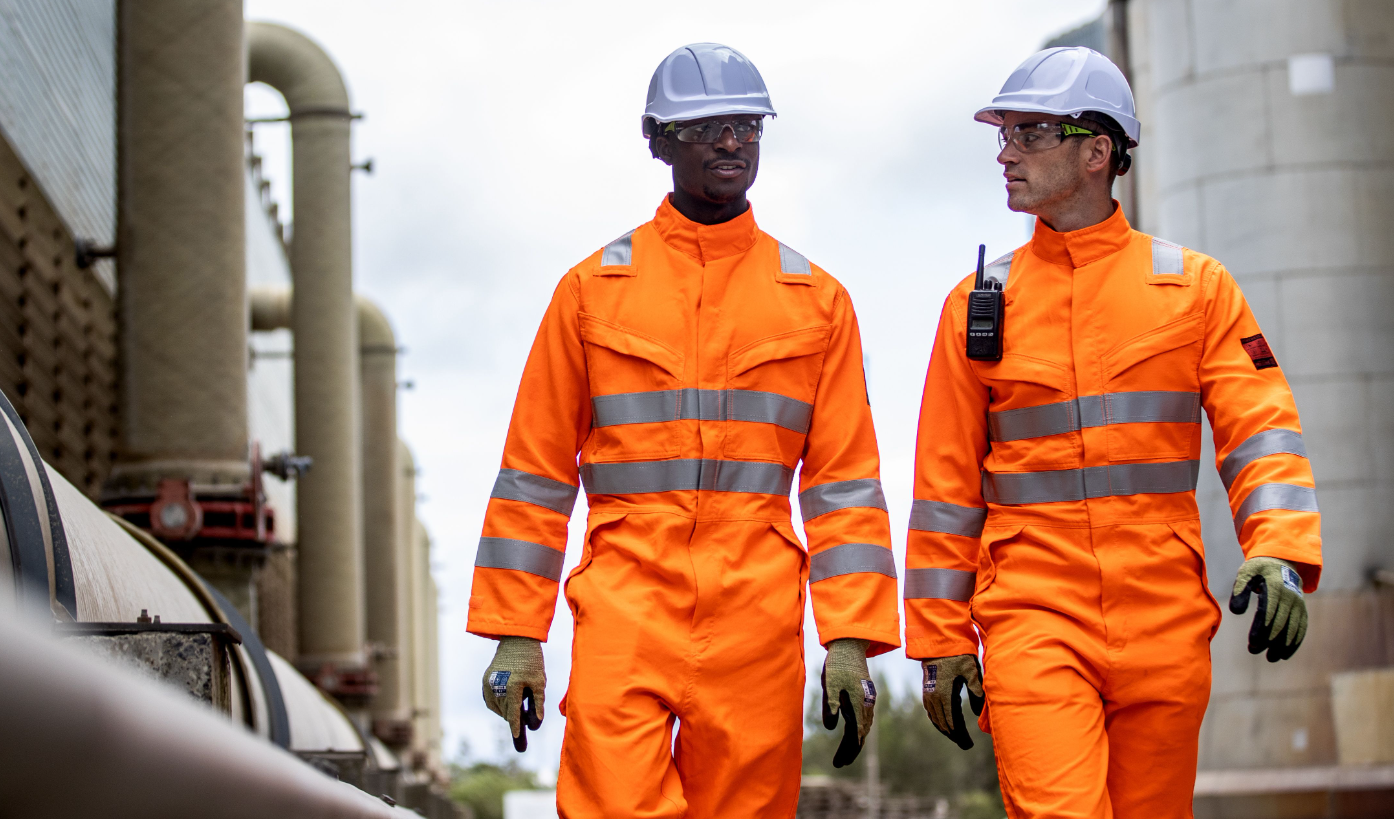 Dos trabajadores frente a un fondo industrial vistiendo ropa naranja de alta visibilidad y cascos blancos. Hay disponible un enlace al casco Endurance Glowtex PG54.