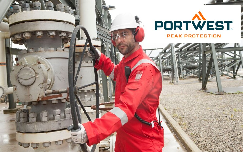 Un trabajador industrial lleva un traje de protección rojo con bandas reflectantes, casco blanco, protección auditiva y gafas de seguridad. Opera un gran volante negro en una planta industrial, presumiblemente una refinería o un patio industrial. Al fondo se ven tuberías y estructuras de acero. El logotipo de la marca Portwest con el eslogan "Peak Protection" se encuentra en la parte superior derecha de la imagen. Se proporciona un enlace a nuestros overoles ignífugos.