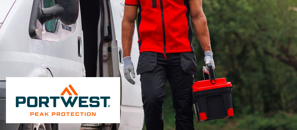 Se puede ver la zona entre los hombros y las pantorrillas de una persona con ropa de trabajo. El hombre lleva un chaleco softshell rojo, una camiseta roja debajo, pantalones negros con bolsillos y guantes blancos. Lleva una caja de herramientas en la mano izquierda. A su izquierda, al fondo, se ve una furgoneta blanca; el resto del fondo está cubierto de árboles caducifolios verdes. El logotipo azul y naranja del fabricante Portwest sobre fondo blanco se encuentra en la esquina inferior izquierda de la imagen.