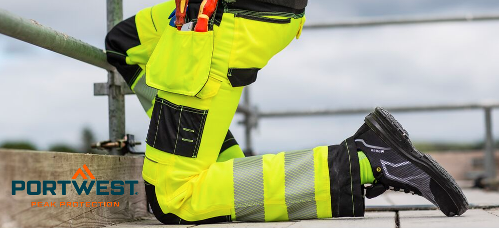 Un obrero de la construcción se arrodilla sobre el suelo de un andamio. Lleva pantalones de trabajo amarillo brillante de alta visibilidad con rodilleras integradas y calzado de seguridad negro. Al fondo se ve un cielo nublado. El logotipo del fabricante, Portwest, en letras azules y naranjas, aparece en la esquina inferior izquierda.
