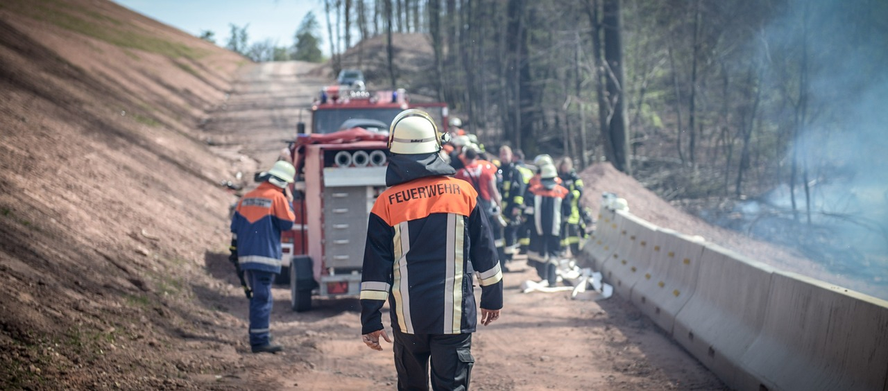 Vista trasera de un bombero en plena marcha. Al fondo se puede ver un camión de bomberos en un camino forestal y un grupo de bomberos trabajando. Se proporciona el enlace al mono ignífugo FR98 FR98 Bizflame FR98 FR98 Bizflame FR.