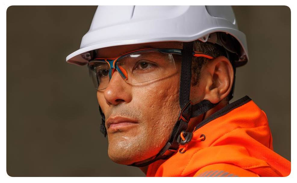 Retrato de un hombre con ropa de alta visibilidad naranja, casco de seguridad blanco y gafas de protección. Hay un enlace a nuestra colección de gafas.