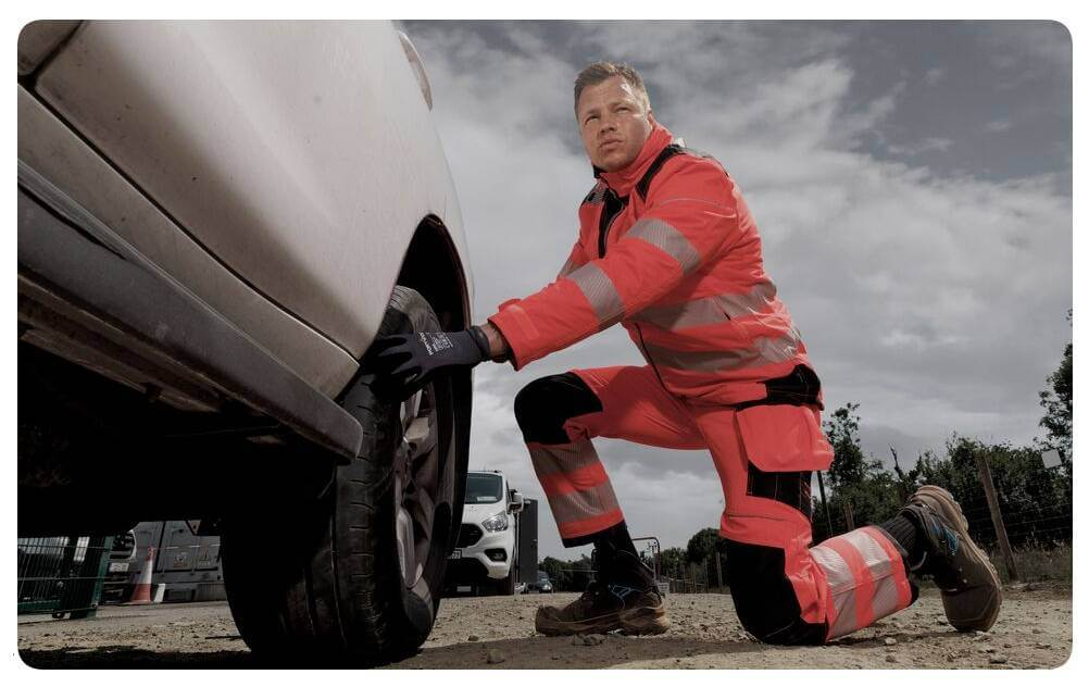 Un hombre con ropa de alta visibilidad naranja está arrodillado delante de un coche blanco y trabaja en el neumático. Al fondo se ve un cielo nublado. Hay un enlace a nuestra colección de protección para rodillas.