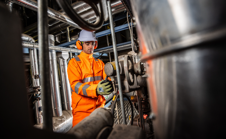Trabajador con casco blanco y protección para los oídos en ropa de trabajo naranja con guantes en un contexto industrial. La imagen está vinculada a la colección de guantes de trabajo.
