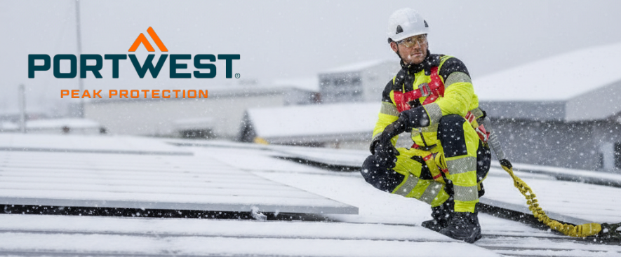 Un trabajador con un mono amarillo de alta visibilidad con bandas reflectantes se arrodilla sobre un tejado nevado junto a un sistema fotovoltaico. Lleva un casco blanco y protecci&oacute;n antica&iacute;das. Se ven m&aacute;s tejados nevados al fondo, y el logotipo del fabricante, Portwest, se ve en azul y naranja en la esquina superior izquierda de la imagen.