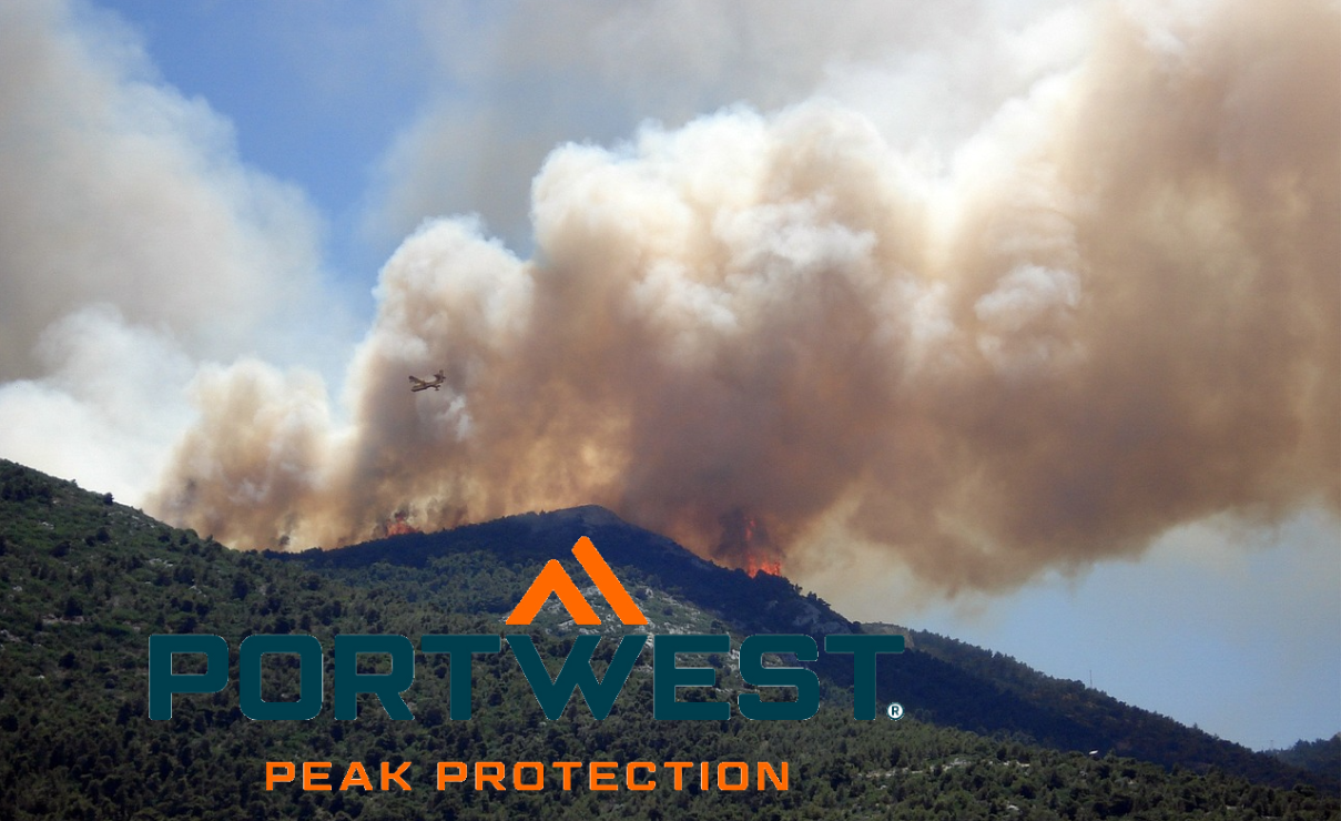 Un incendio forestal arde en la ladera de una montaña verde, con densas nubes de humo elevándose hacia el cielo. Un avión de bomberos se acerca, arrojando agua o agente extintor sobre las llamas. El logotipo de la marca Portwest se ve en primer plano; el eslogan "Peak Protection" se encuentra debajo. Se incluye un enlace a nuestra selección de ropa para bomberos.