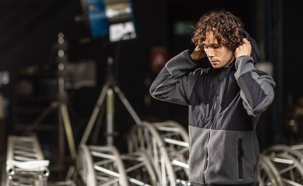 Hombre de cabello rizado con una chaqueta de forro polar gris con capucha y detalles negros, de pie en un entorno industrial con estructuras metálicas y focos. Enlace a nuestra colección de forros polares.