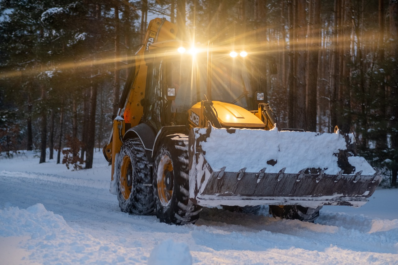 Tractor amarillo con una pala grande en un sendero forestal nevado. El crepúsculo va cayendo lentamente y el vehículo tiene una luz encendida.